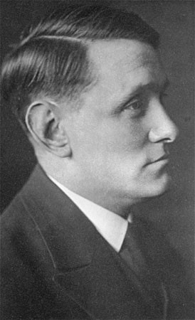A man with light skin tone and dark hair faces the right. He is wearing a dark suit and tie.