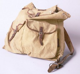 Tan cloth backpack with drawstring top and flap cover. The front features two pockets with brown leather closures. One brown leather shoulder strap and buckle is shown on the right side.